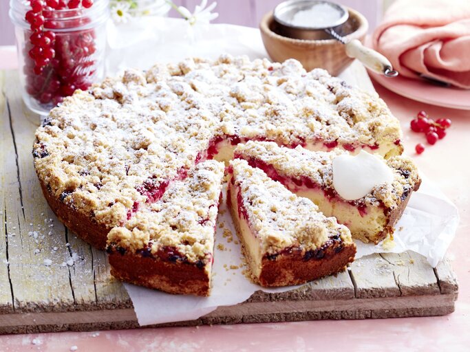 Johannisbeer-Käsekuchen mit Streuseln auf einem Holzbrett serviert, ein Stück angeschnitten, daneben ein Glas mit roten Johannisbeeren und Gänseblümchen. | © EAT CLUB/Isabell Triemer
