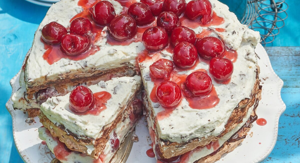 Angeschnittene Schwarzwälder Kirsch-Baisertorte mit Kirschen oben drauf auf blauem Untergrund. 