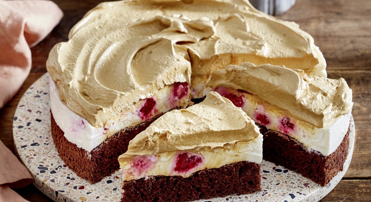 Dalgona-Himbeer-Cheesecake auf einem Tortenteller auf hölzernem Hintergrund. Links davon ein rosarotes Geschirrtuch. 