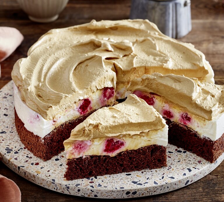 Dalgona-Himbeer-Cheesecake auf einem Tortenteller auf hölzernem Hintergrund. Links davon ein rosarotes Geschirrtuch.  | © EAT CLUB/Isabell Triemer