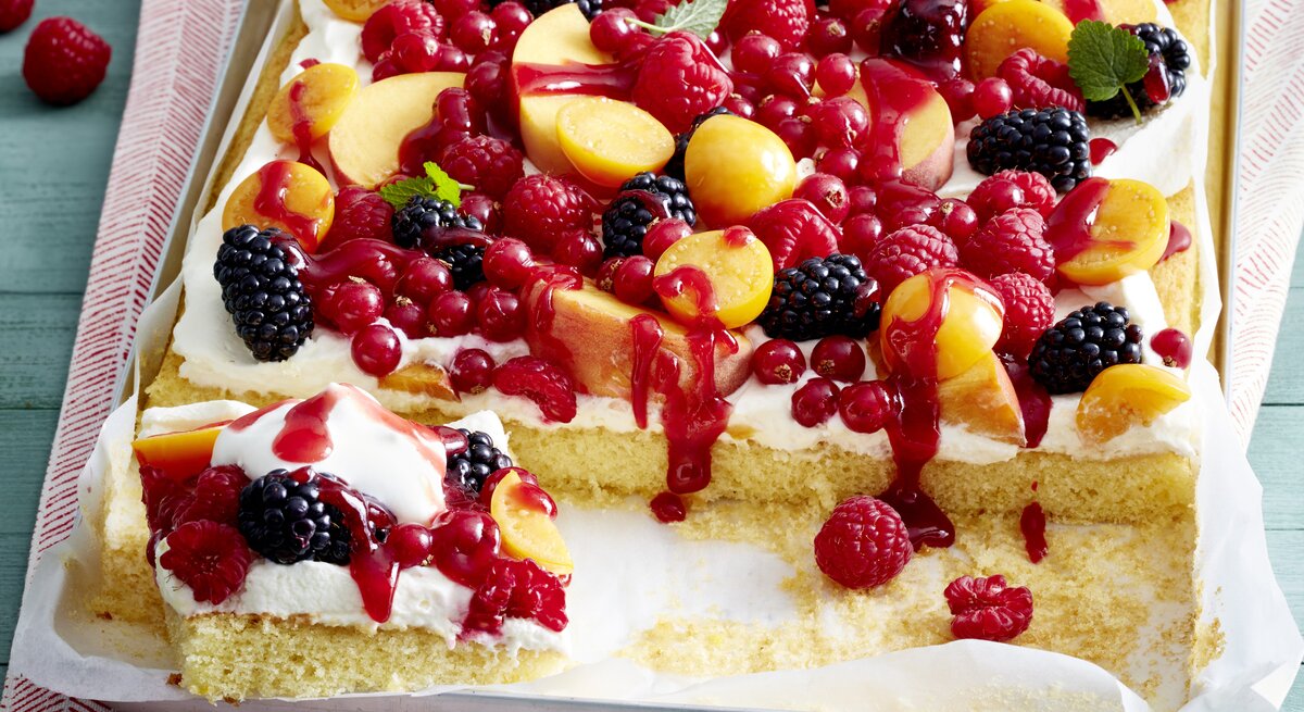 Tutti Frutti Schnitten auf einem weißen Blech auf einem rot-weißen-Geschirrtuch vor grünem Hintergrund. Hinten auf dem Foto Himbeeren.