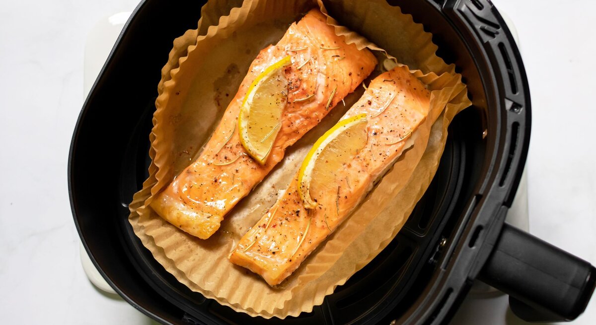 Zwei goldbraun gebackene Lachsfilets mit Zitronenscheiben und Gewürzen liegen auf Backpapier in einem schwarzen Airfryer.