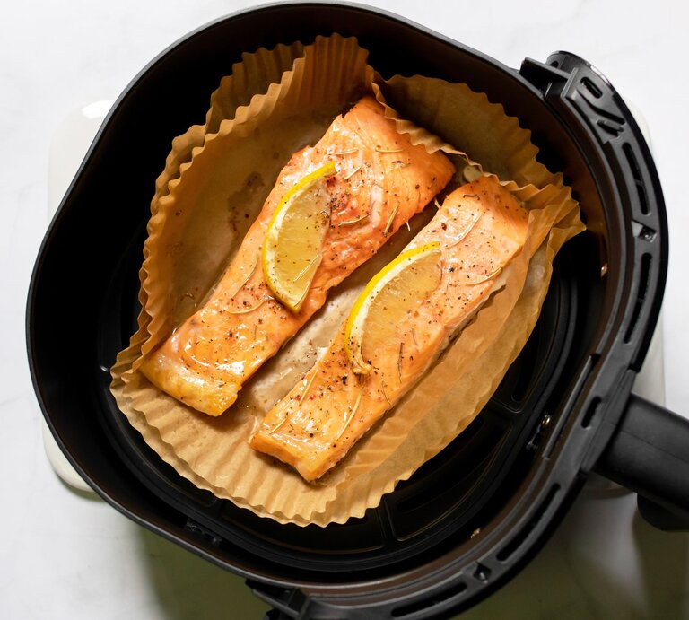 Zwei goldbraun gebackene Lachsfilets mit Zitronenscheiben und Gewürzen liegen auf Backpapier in einem schwarzen Airfryer. | © Shutterstock/PJjaruwan