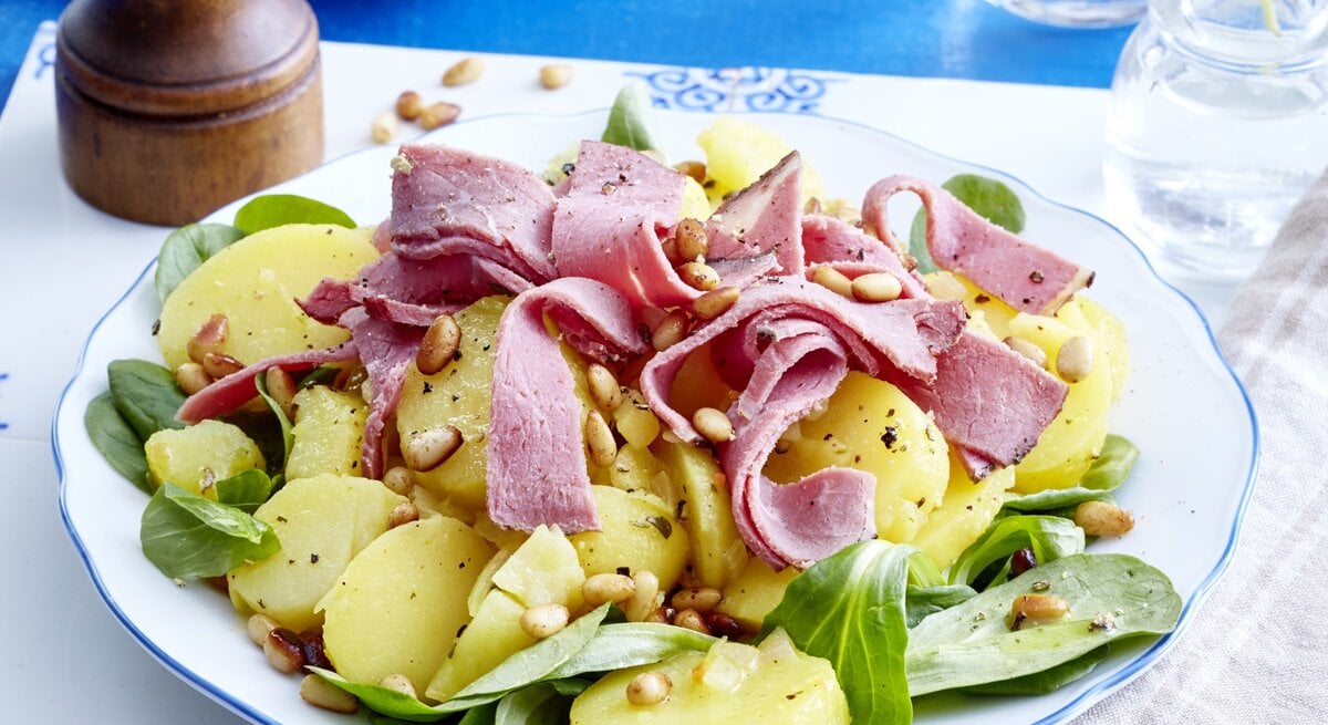 Kartoffelsalat mit Roastbeef auf einem weißen Teller auf einem weißen Tischuntersetzer auf einem blauen Tisch. Rechts ein weiß/graues Geschirrtuch und links eine Pfeffermühle.