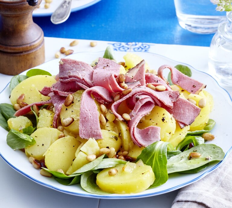 Kartoffelsalat mit Roastbeef auf einem weißen Teller auf einem weißen Tischuntersetzer auf einem blauen Tisch. Rechts ein weiß/graues Geschirrtuch und links eine Pfeffermühle. | © EAT CLUB/Isabell Triemer