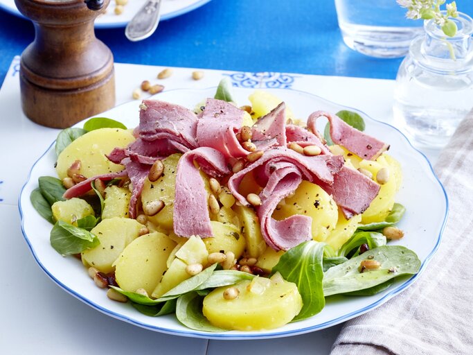 Kartoffelsalat mit Roastbeef auf einem weißen Teller auf einem weißen Tischuntersetzer auf einem blauen Tisch. Rechts ein weiß/graues Geschirrtuch und links eine Pfeffermühle. | © EAT CLUB/Isabell Triemer