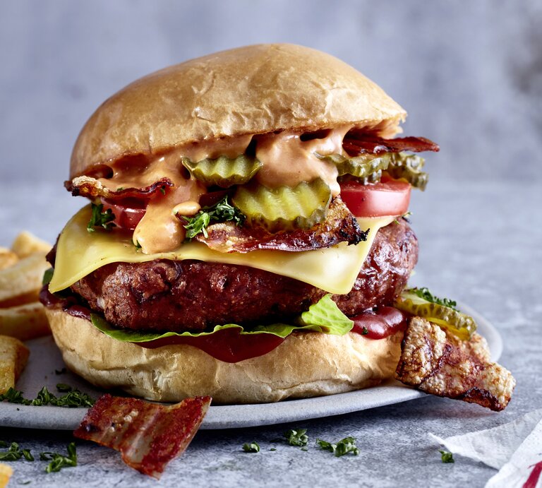 Kompromisslos guter cheesy Böhnchenburger auf einem Teller mit Pommes angerichtet. Der Hintergrund ist grau. | © EAT CLUB/Isabell Triemer
