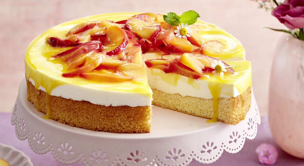 Nektarinen-Joghurt-Torte auf einem weißen Tortenteller vor rosa und lila Hintergrund. Links kleine Teller und rechts auf dem Foto Blumen in einer Vase.