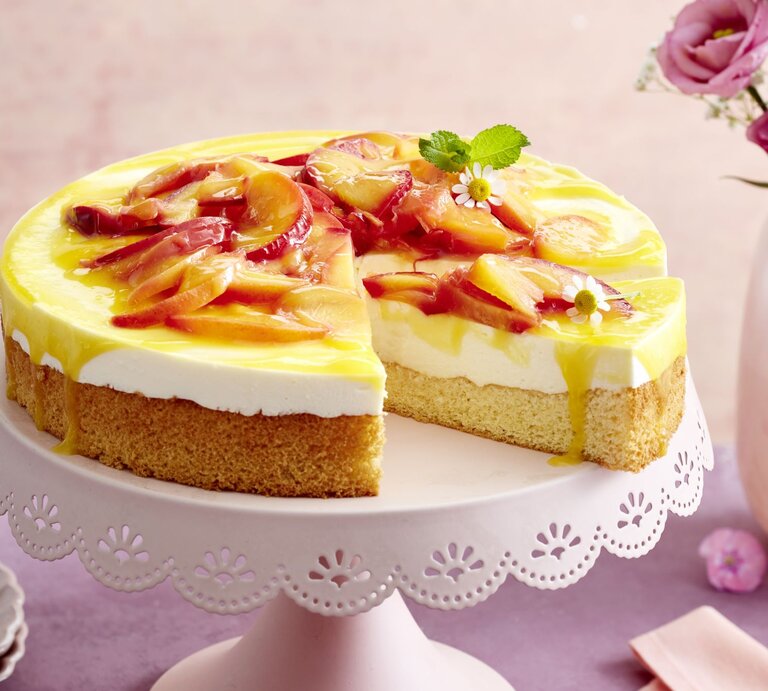 Nektarinen-Joghurt-Torte auf einem weißen Tortenteller vor rosa und lila Hintergrund. Links kleine Teller und rechts auf dem Foto Blumen in einer Vase. | © EAT CLUB/Isabell Triemer