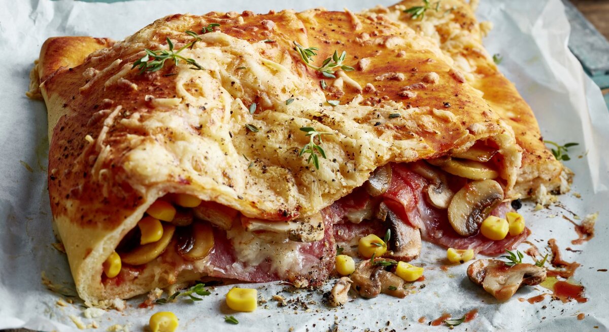 Pizza Calzone mit Mais, Pilzen und Salami auf weißem Backpapier vor hölzernem, leicht blau bemaltem Hintergrund.