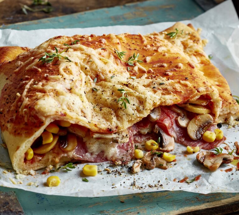 Pizza Calzone mit Mais, Pilzen und Salami auf weißem Backpapier vor hölzernem, leicht blau bemaltem Hintergrund. | © EAT CLUB/Isabell Triemer