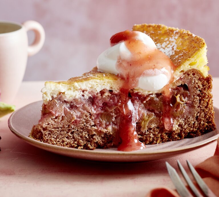Ein Stück Rhabarber-Torte mit Schmand auf einem rosa Teller vor rosa Hintergrund. Hinten auf dem Foto eine Tasse. | © EAT CLUB/Isabell Triemer