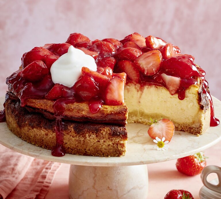 Erdbeer-Käsekuchen mit Sahne auf einem beigen Tortenteller vor rosa Hintergrund, darauf Erdbeeren und eine Tasse Kaffee verteilt.  | © EAT CLUB/Isabell Triemer