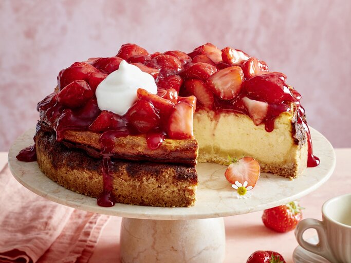 Erdbeer-Käsekuchen mit Sahne auf einem beigen Tortenteller vor rosa Hintergrund, darauf Erdbeeren und eine Tasse Kaffee verteilt.  | © EAT CLUB/Isabell Triemer
