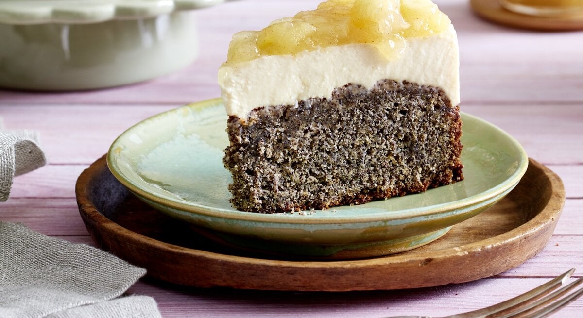 Ein Stück Joghurtcreme-Torte auf einem grünen Teller auf einem braunen Unterteller. Der Hintergrund ist lila. Rechts unten auf dem Foto eine Gabel, links unten ein Geschirrtuch. 