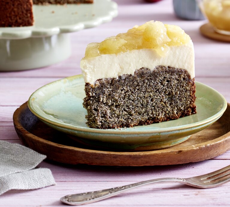 Ein Stück Joghurtcreme-Torte auf einem grünen Teller auf einem braunen Unterteller. Der Hintergrund ist lila. Rechts unten auf dem Foto eine Gabel, links unten ein Geschirrtuch.  | © EAT CLUB/Isabell Triemer