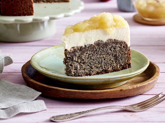 Ein Stück Joghurtcreme-Torte auf einem grünen Teller auf einem braunen Unterteller. Der Hintergrund ist lila. Rechts unten auf dem Foto eine Gabel, links unten ein Geschirrtuch.  | © EAT CLUB/Isabell Triemer