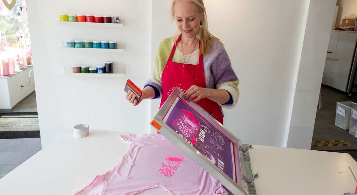 Lächelnde Frau mit roter Schürze bedruckt ein T-Shirt per Siebdruck, im Hintergrund bunte Farbdosen an weißer Wand.