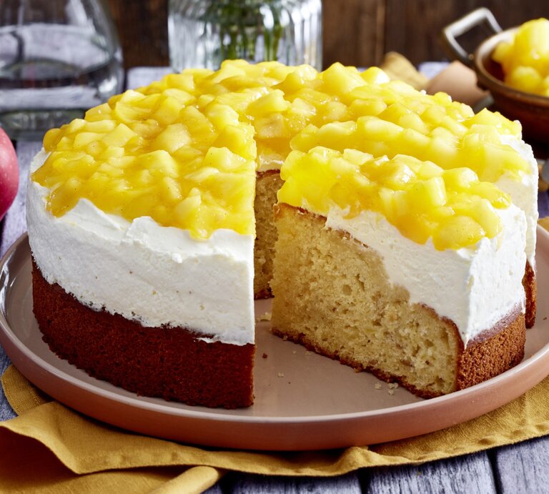 Apfel-Schmandkuchen auf einem Teller auf einem gelben Geschirrtuch. Dahinter Äpfel, Geschirr, Besteck und Wasser. | © EAT CLUB/Isabell Triemer