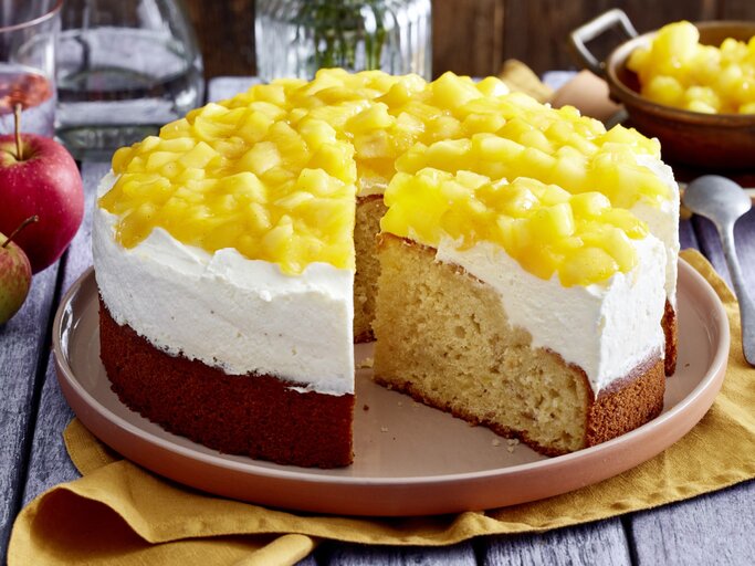 Apfel-Schmandkuchen auf einem Teller auf einem gelben Geschirrtuch. Dahinter Äpfel, Geschirr, Besteck und Wasser. | © EAT CLUB/Isabell Triemer