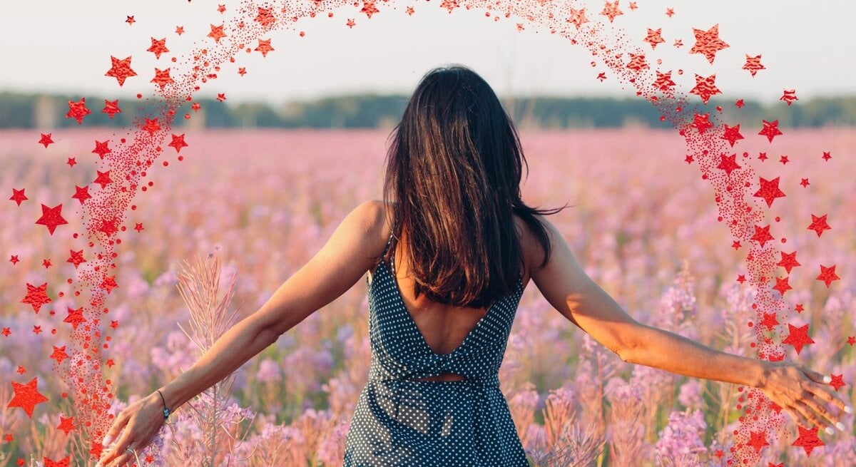 Eine Frau mit dunkelbraunem Haar in einem ärmellosen, gepunkteten Kleid steht mit dem Rücken zur Kamera in einem weiten Feld voller rosa-lila Wildblumen. Ihre Arme sind seitlich ausgestreckt, während sie die Blumen berührt. Um sie herum ist ein digital eingefügter Halbkreis aus roten Sternen zu sehen, der wie ein magischer Funkenbogen wirkt. Der Himmel ist hell und klar, im Hintergrund ist eine Baumlinie zu erkennen.