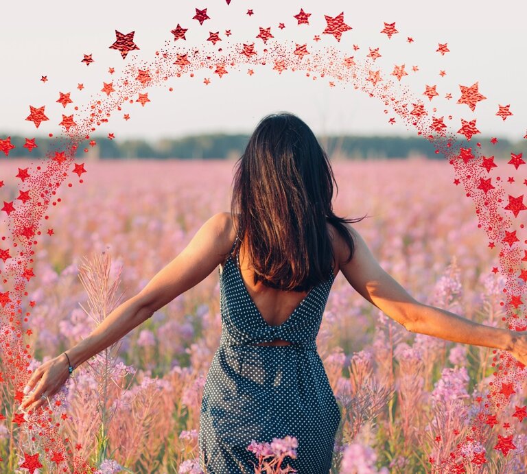 Eine Frau mit dunkelbraunem Haar in einem ärmellosen, gepunkteten Kleid steht mit dem Rücken zur Kamera in einem weiten Feld voller rosa-lila Wildblumen. Ihre Arme sind seitlich ausgestreckt, während sie die Blumen berührt. Um sie herum ist ein digital eingefügter Halbkreis aus roten Sternen zu sehen, der wie ein magischer Funkenbogen wirkt. Der Himmel ist hell und klar, im Hintergrund ist eine Baumlinie zu erkennen. | © NassyArt und primipil via Canva.com [M]