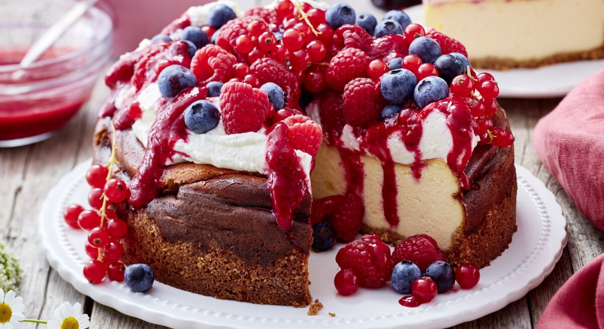 Ein angeschnittener Beeren-Käsekuchen mit Schlagsahne und verschiedenen frischen Beeren darauf auf einem weißen Teller und heller, verschwommener Hintergrund. 