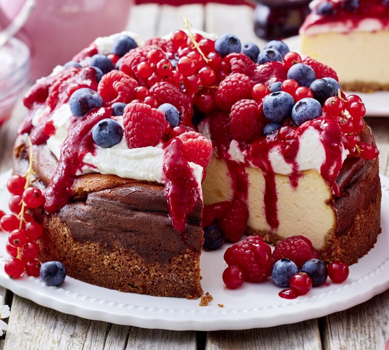 Ein angeschnittener Beeren-Käsekuchen mit Schlagsahne und verschiedenen frischen Beeren darauf auf einem weißen Teller und heller, verschwommener Hintergrund.  | © EAT CLUB / Isabell Triemer 