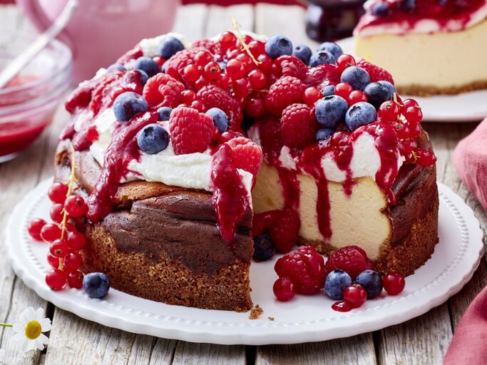 Ein angeschnittener Beeren-Käsekuchen mit Schlagsahne und verschiedenen frischen Beeren darauf auf einem weißen Teller und heller, verschwommener Hintergrund.  | © EAT CLUB / Isabell Triemer 