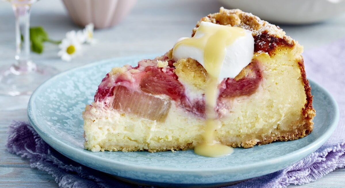 Rhabarber-Käsekuchen auf einem blauen Hintergrund. Hinten Gläser und Gänseblümchen verteilt.
