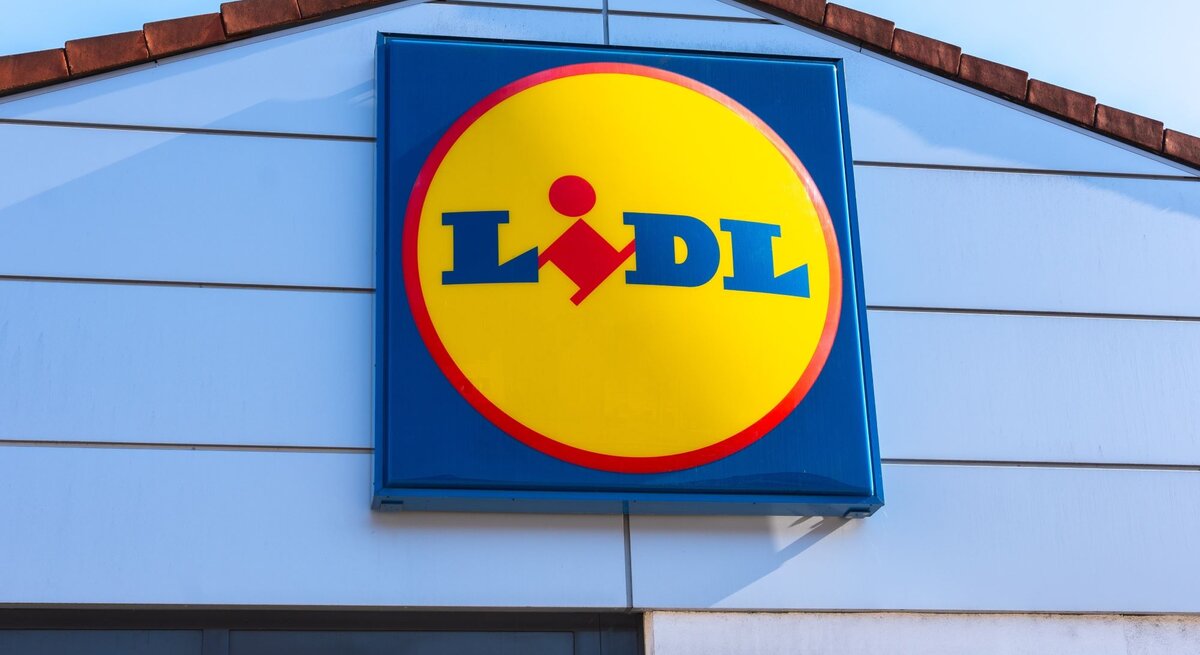 Lidl-Logo in gelber Kreisform mit rotem Rand an der hellblauen Fassade eines Filialgebäudes unter klarem Himmel.