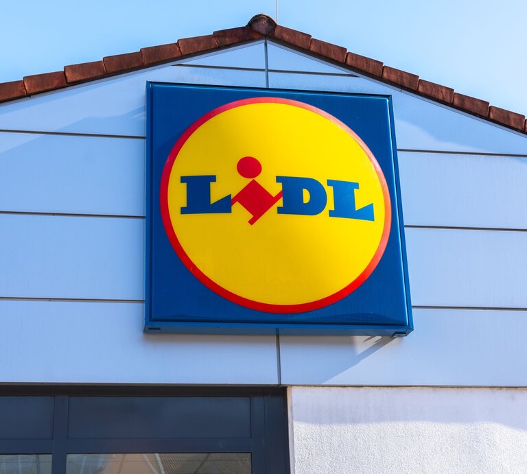 Lidl-Logo in gelber Kreisform mit rotem Rand an der hellblauen Fassade eines Filialgebäudes unter klarem Himmel. | © Shutterstock/ Elena Ebinger