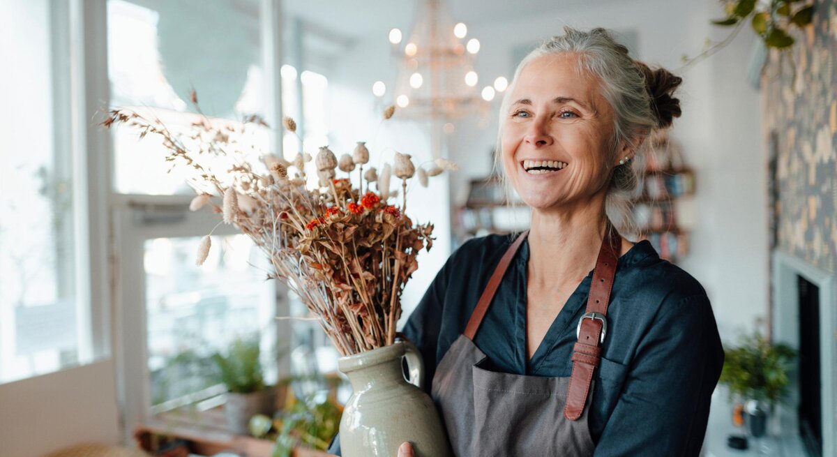 Ältere Frau arbeitet nebenbei in einem Blumenladen und hält eine Vase mit Blumen, dabei lächelt sie. 