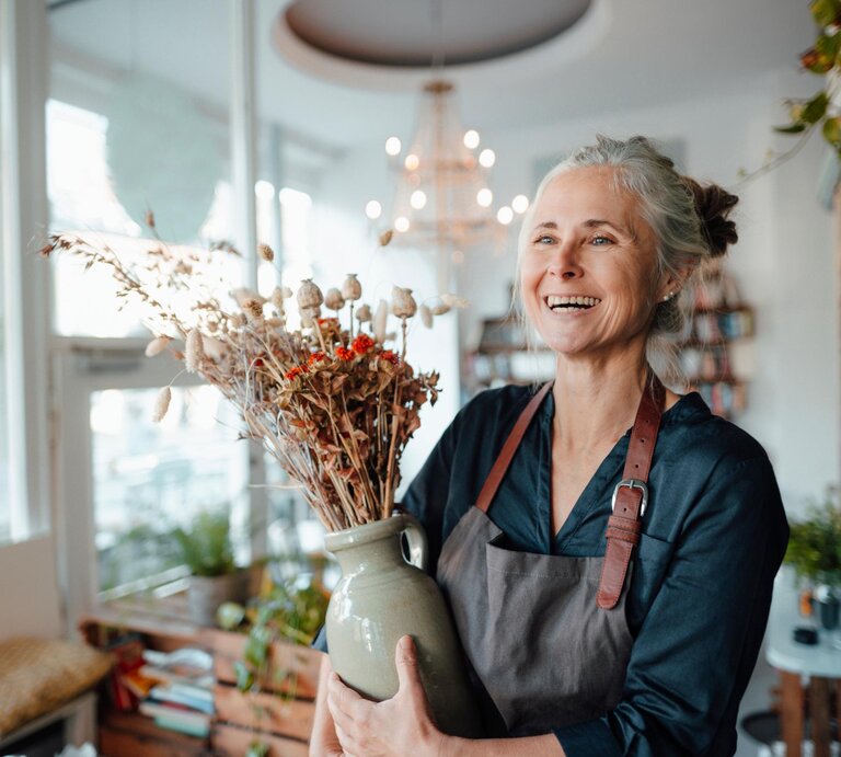 Ältere Frau arbeitet nebenbei in einem Blumenladen und hält eine Vase mit Blumen, dabei lächelt sie.  | © Getty Images