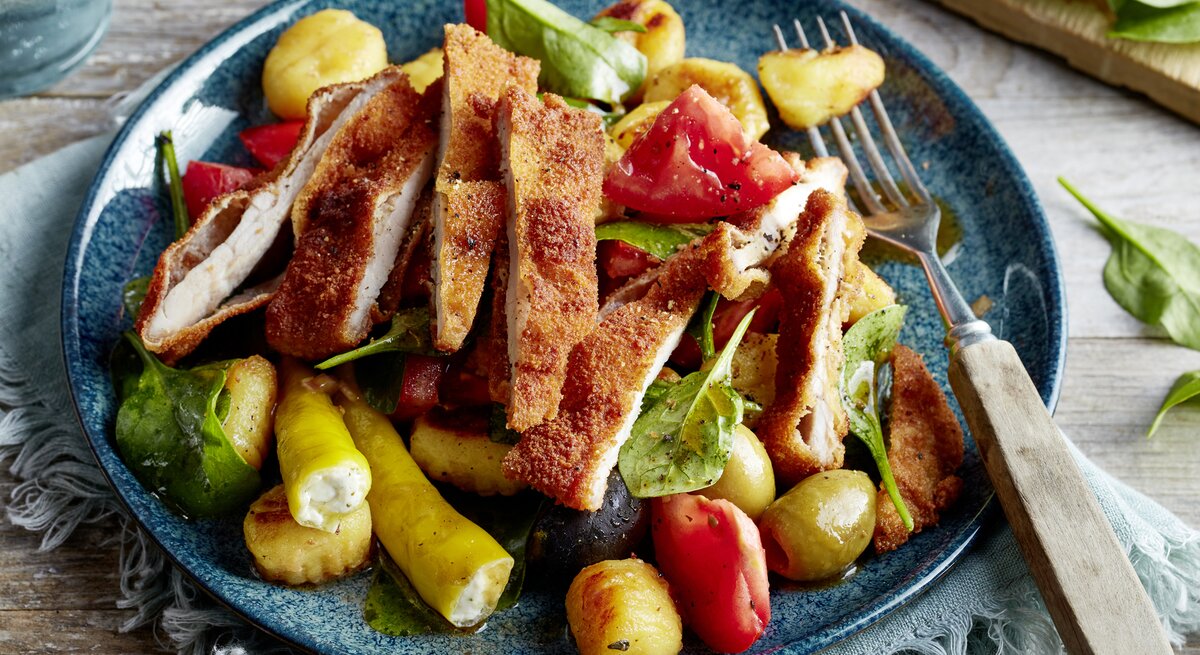 Antipasti-Schnitzel-Salat auf einem blauen Teller auf einem hellblauen Geschirrtuch auf hölzernem Hintergrund.