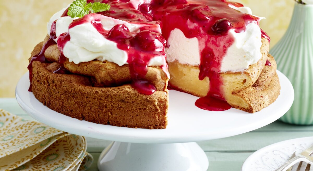 Rote-Grütze-Torte mit Puddingboden auf einem weißen Tortenteller auf grünem Hintergrund.