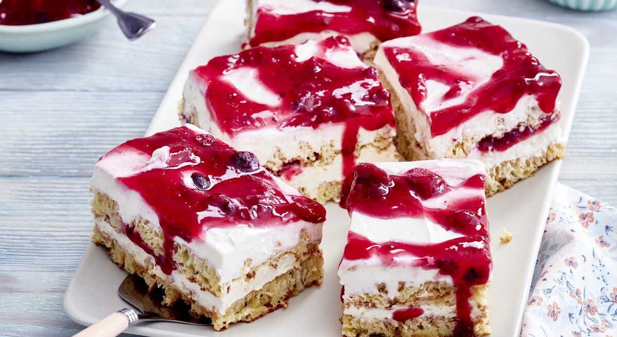 Rote-Grütze-Tiramisu-Schnitten auf einem weißen Teller auf grauem Hintergrund. Links oben eine Schüssel mit Roter Grütze.