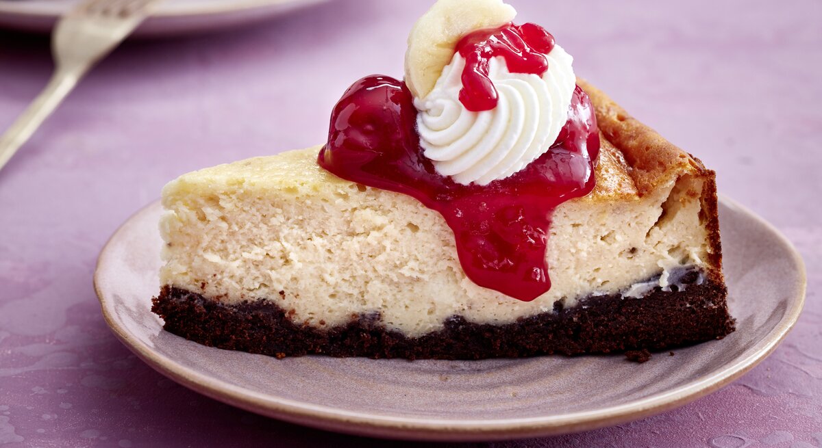 Ein Stück KiBa-Cheesecake auf einem Teller auf rosa Hintergrund. Vorne auf dem Foto eine Gabel.