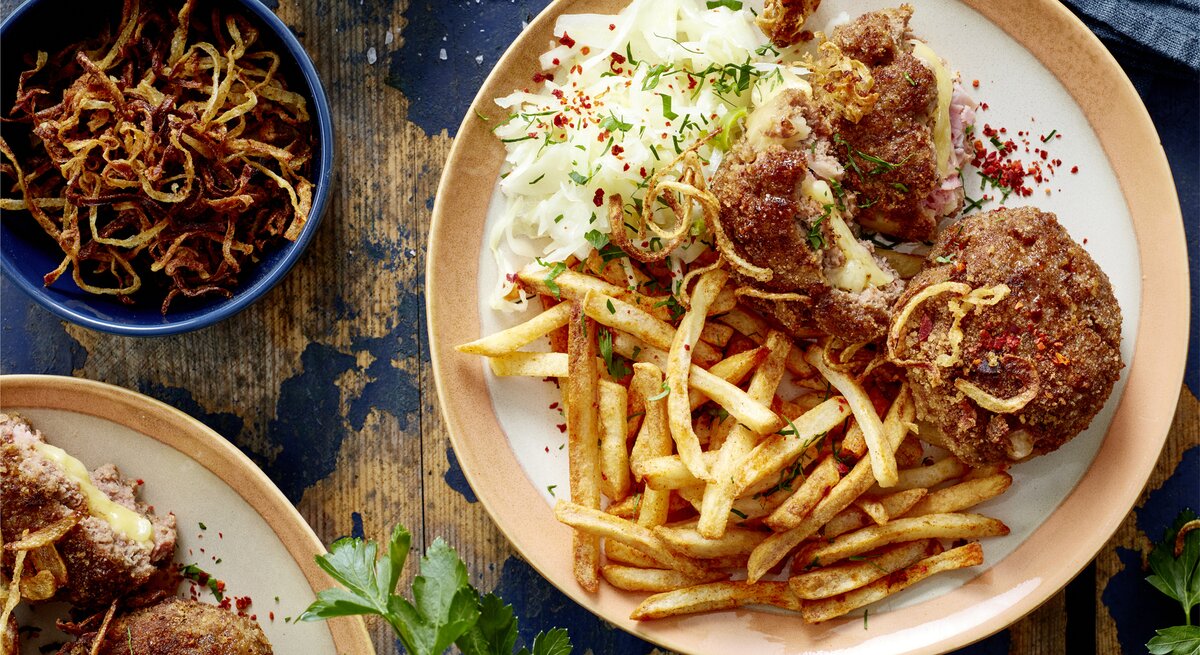  Cordon-bleu-Frikadellen mit Pommes auf einem Teller auf hölzernem Hintergrund. Links davon Röstzwiebeln, Salz und weitere Frikadellen.