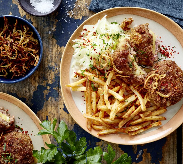  Cordon-bleu-Frikadellen mit Pommes auf einem Teller auf hölzernem Hintergrund. Links davon Röstzwiebeln, Salz und weitere Frikadellen. | © EAT CLUB / Isabell Triemer