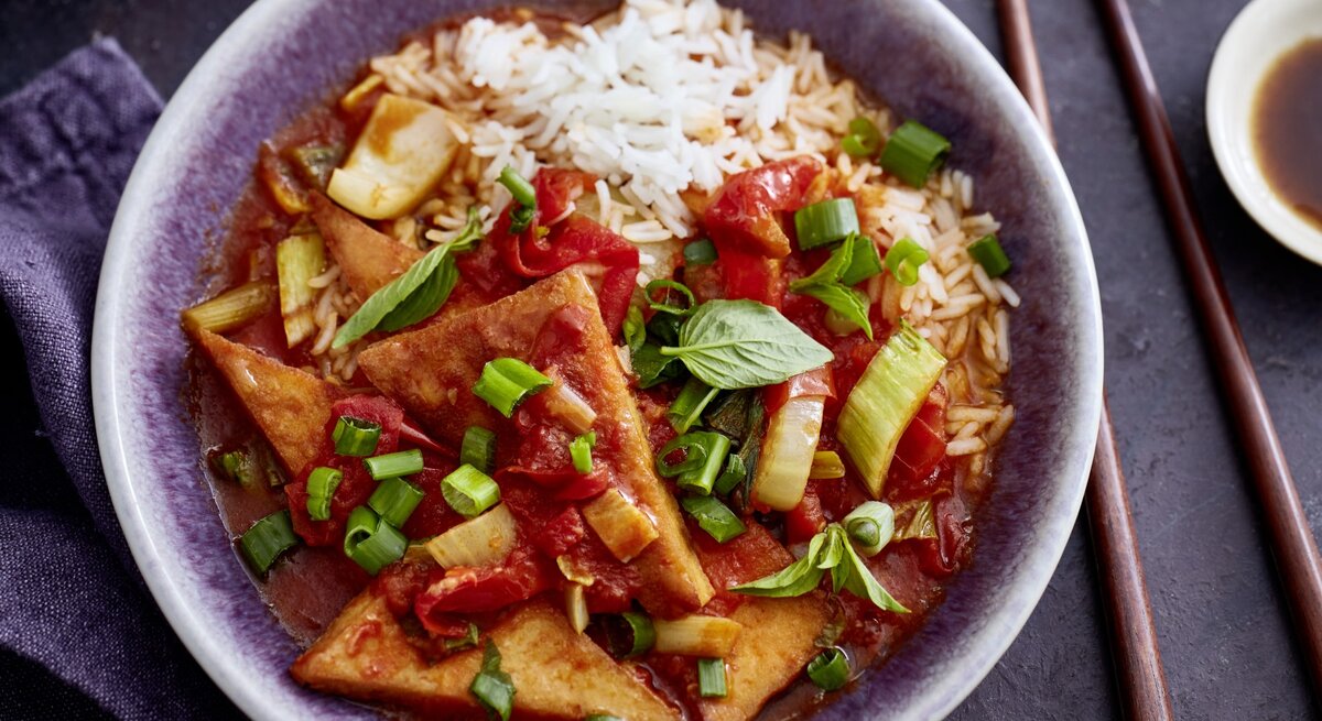 Tofu süßsauer mit Pak Choi und Paprika in einer grauen Schüssel auf grauem Hintergrund. Rechts von der Bowl sind Stäbchen. 