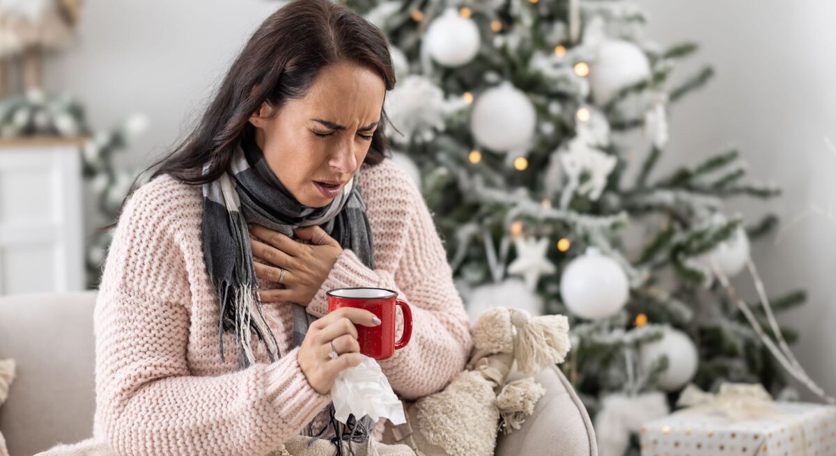 Eine dunkelhaarige Frau in einem rosa Strickpullover sitzt krank auf dem Sofa, hält eine Tasse Tee und presst die Hand an die Brust.