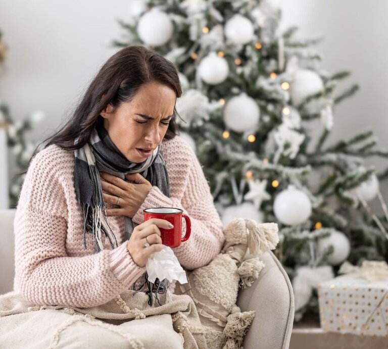 Eine dunkelhaarige Frau in einem rosa Strickpullover sitzt krank auf dem Sofa, hält eine Tasse Tee und presst die Hand an die Brust. | © GettyImages/SimpleImages