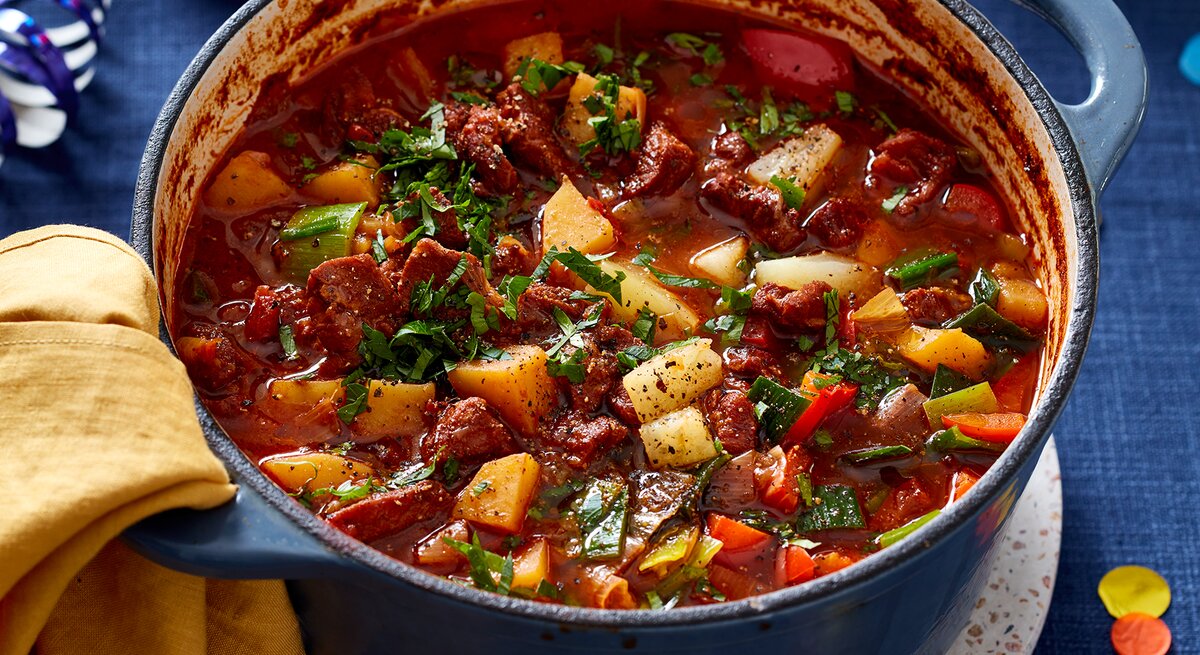 Ofen-Gulaschsuppe in einem blauen Topf auf blauem Hintergrund. Links auf dem Foto ein gelbes Geschirrtuch.
