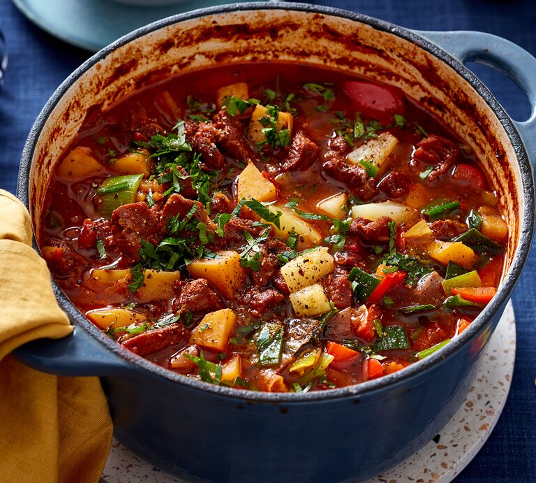 Ofen-Gulaschsuppe in einem blauen Topf auf blauem Hintergrund. Links auf dem Foto ein gelbes Geschirrtuch. | © EAT CLUB/Isabell Triemer