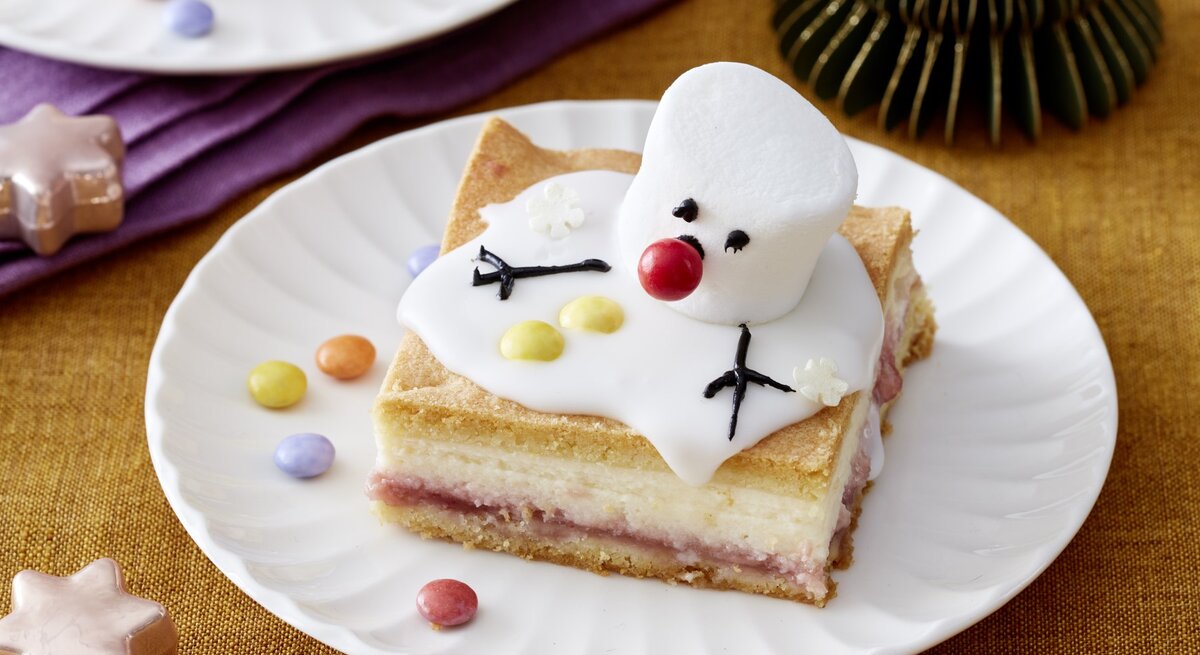 Ein Stück Schneemann-Käsekuchen auf einem weißen Teller. Auf dem Hintergrund Zimtsterne verteilt.