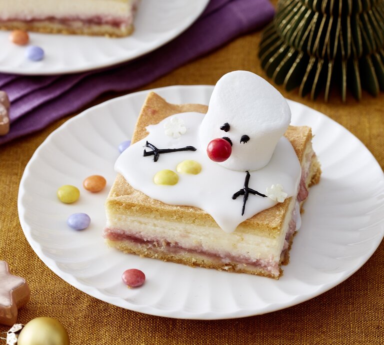 Ein Stück Schneemann-Käsekuchen auf einem weißen Teller. Auf dem Hintergrund Zimtsterne verteilt. | © EAT CLUB / Isabell Triemer