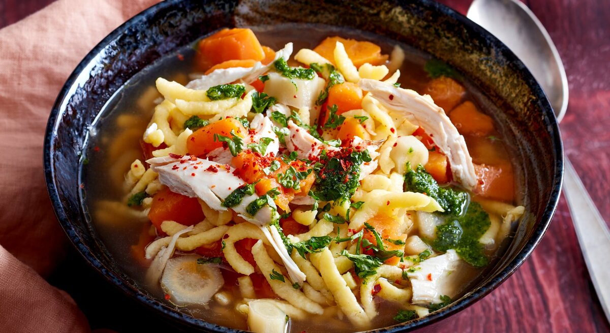 Hühnertopf mit Spätzle in einer dunkelblauen Schüssel auf hölzernem Hintergrund. Links ein rosa Geschirrtuch, rechts ein Löffel.