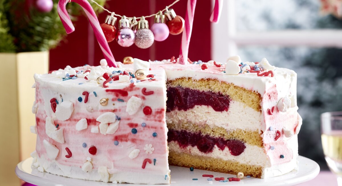 Zuckerstangen-Torte auf einem weißen Tortenteller auf einer rosa Tischdecke dekoriert mit kleinen Weihnachtskugeln. Links hinten auf dem Foto ein kleiner Tannenbaum.