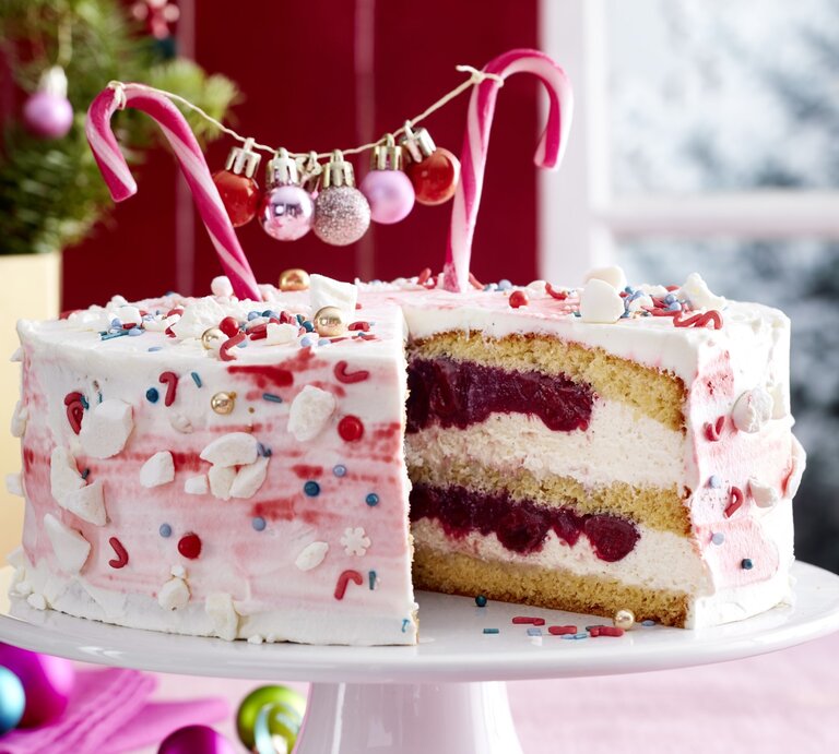 Zuckerstangen-Torte auf einem weißen Tortenteller auf einer rosa Tischdecke dekoriert mit kleinen Weihnachtskugeln. Links hinten auf dem Foto ein kleiner Tannenbaum. | © EAT CLUB / Isabell Triemer